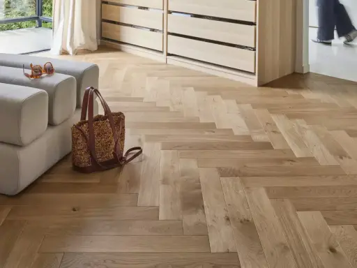Un parquet en bois clair au calepinage en chevrons, avec un canapé, un sac et des lunettes posés dessus.