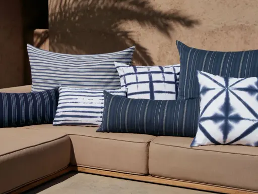 Plusieurs coussins bleus et blancs, à rayures et motifs tie-dye, sont disposés sur un canapé marron. Idée pour comment mettre un plaid sur un canapé.