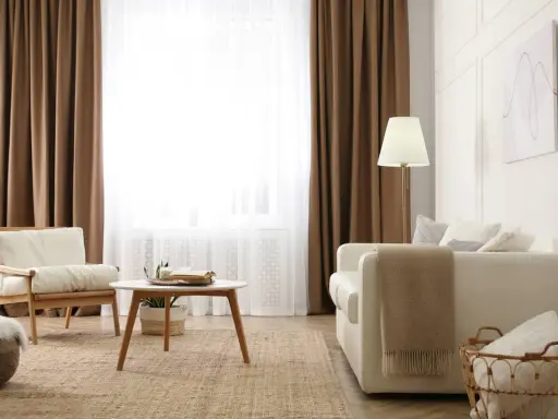 Salon cosy avec 1 ou 2 rideaux par fenêtre, fauteuil en bois, canapé blanc, table basse et tapis en jute.