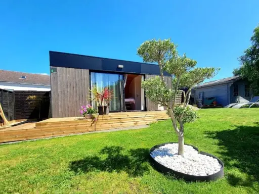 Un charmant pavillon maison moderne avec terrasse en bois, entouré d'un jardin verdoyant et d'un petit olivier.