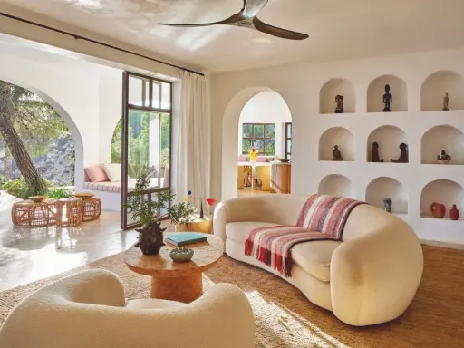 Comment habiller un mur blanc salon : des niches décorées d'objets ethniques et de poteries, un canapé incurvé, des fauteuils bouclette, et une vue sur un jardin luxuriant.