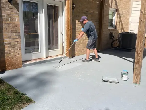 Un homme applique une peinture sur mur béton extérieur avec un rouleau. Le sol gris est en cours de rénovation.