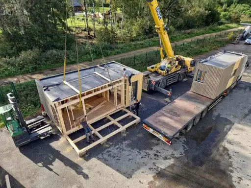 Une grue soulève un module de construction pour des maisons modulaires. Un chariot élévateur et un camion plateau sont également présents.