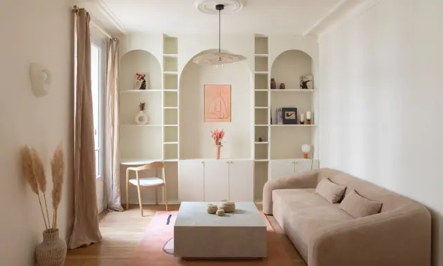 Un coin bureau discret dans un salon cosy, avec une étagère murale arche, un canapé beige et un tapis rose.