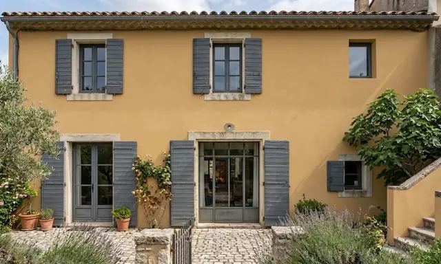 Façade d'une maison provençale à l'enduit projeté ocre, avec volets gris et végétation luxuriante.