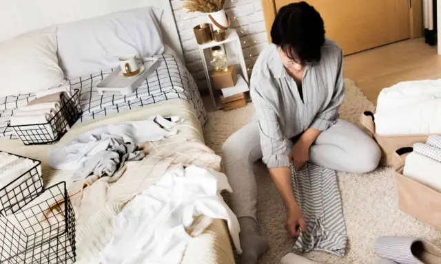 Une femme plie du linge sur un tapis, à côté d'un lit défait. Elle apprend comment ranger sa chambre adulte efficacement.