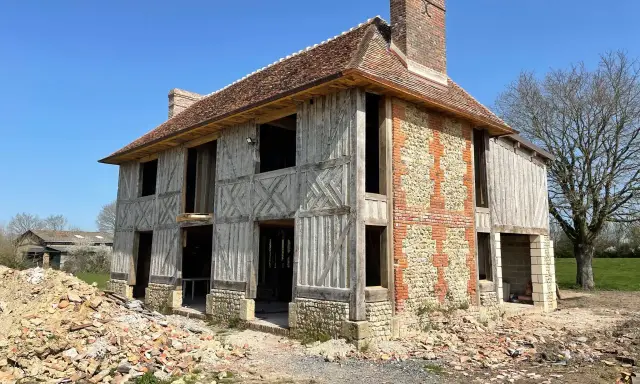 Construction d'une maison normande traditionnelle, avec colombages apparents et murs en pierre et brique. Le chantier est en cours.