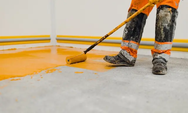 Un ouvrier en combinaison orange et grise, les bottes sales, utilise un rouleau pour peindre le sol d'un garage en jaune vif.