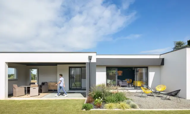Une jeune personne traverse la pelouse devant une petite maison moderne avec une terrasse aménagée et un jardin de gravier.