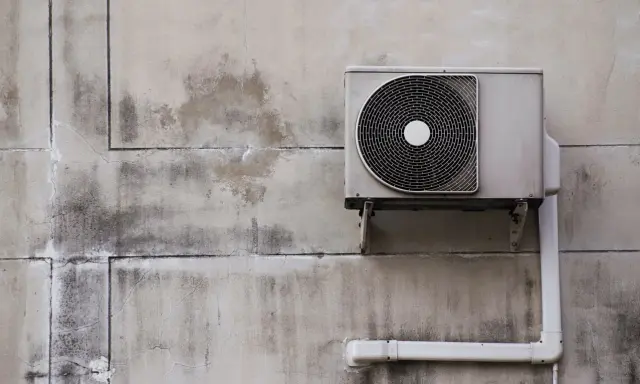 Unité extérieure d'une pompe à chaleur fixée sur un mur en béton. L'isolation est visible sur le tuyau blanc.