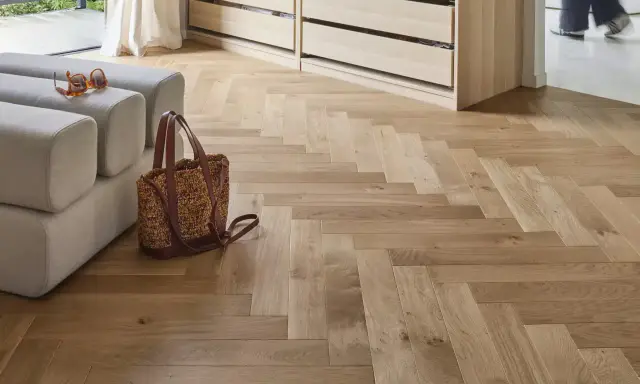 Un parquet en bois clair au calepinage en chevrons, avec un canapé, un sac et des lunettes posés dessus.