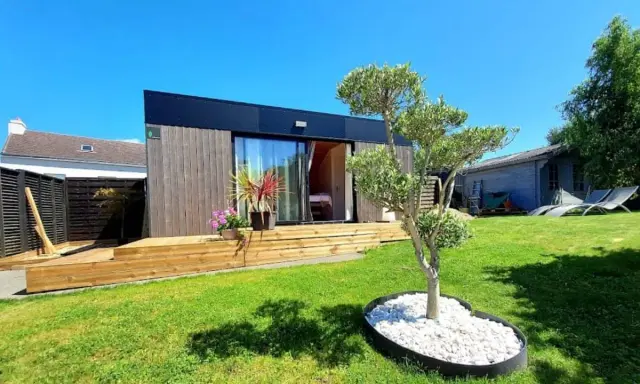 Un charmant pavillon maison moderne avec terrasse en bois, entouré d'un jardin verdoyant et d'un petit olivier.