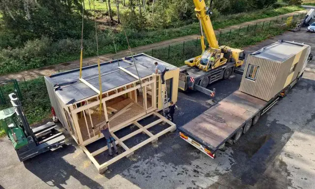 Une grue soulève un module de construction pour des maisons modulaires. Un chariot élévateur et un camion plateau sont également présents.