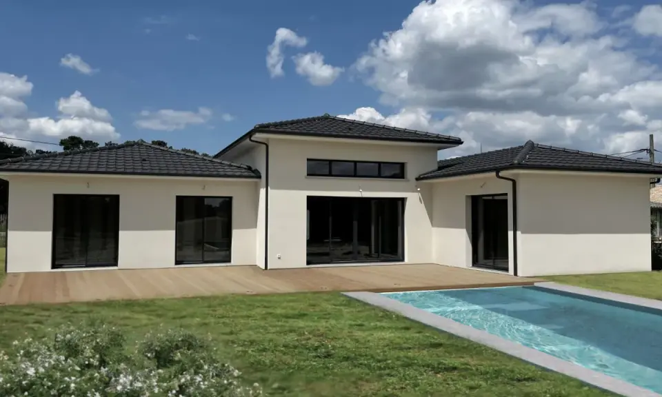 Une maison du sud moderne avec piscine, terrasse en bois et grand jardin sous un ciel bleu nuageux.