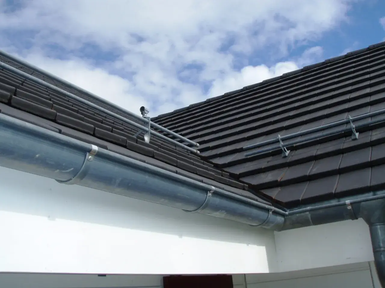 Le chêneau de cette maison, en zinc, est bien fixé sous le toit de tuiles sombres. Le ciel bleu et nuageux promet une belle journée.