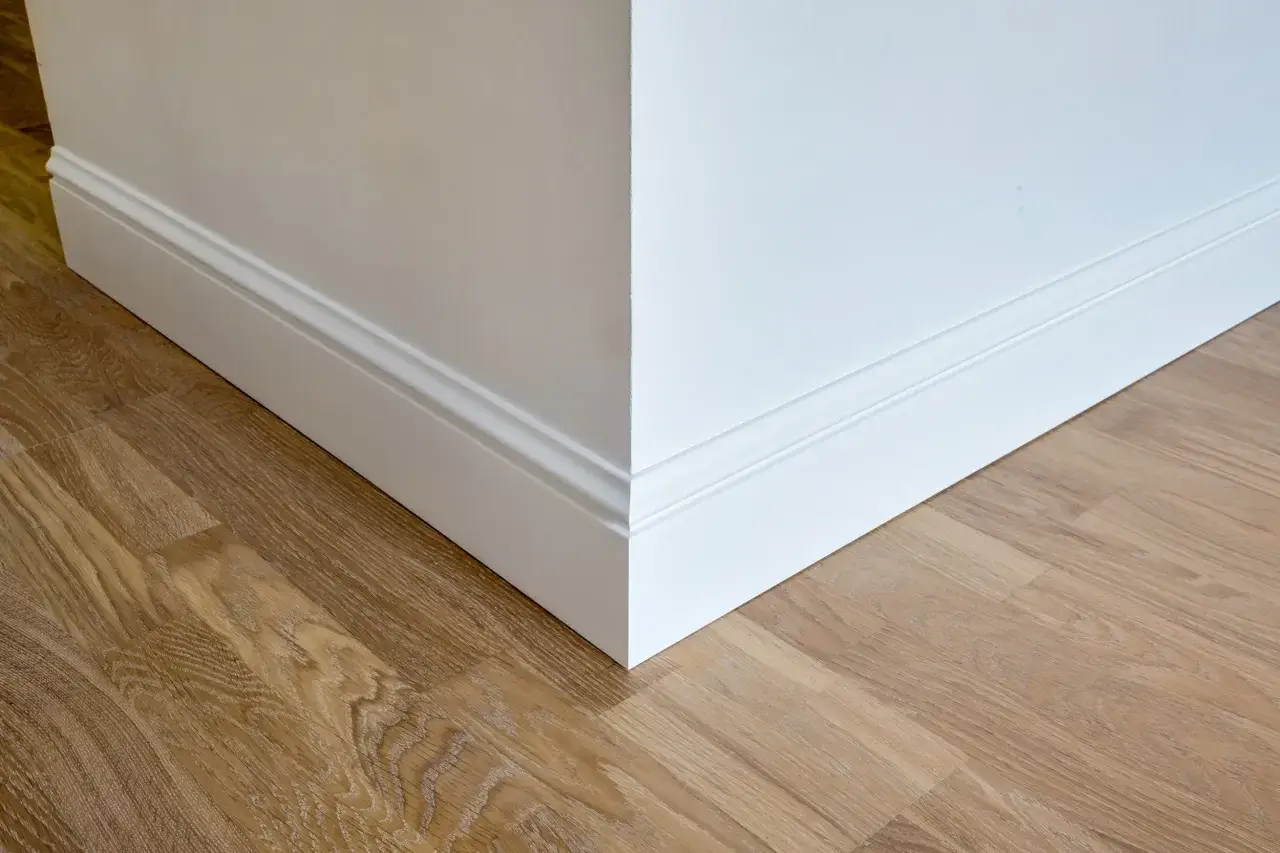 Un coin de mur blanc avec des plinthes moulurées et un parquet en bois clair. Ces plinthes servent à protéger les murs et à cacher les imperfections.