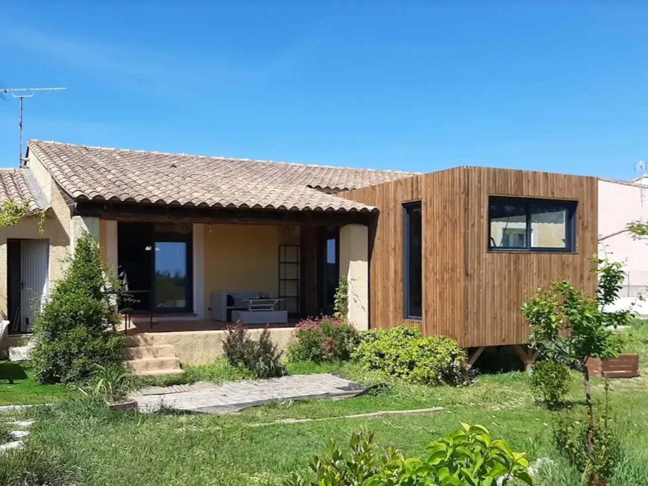 Agrandissement maison ossature bois, ajoutant une extension moderne en bois à une habitation existante. Le contraste des matériaux est saisissant.