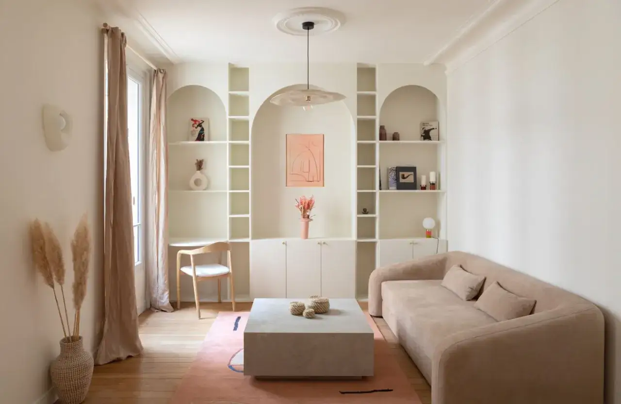 Un coin bureau discret dans un salon cosy, avec une étagère murale arche, un canapé beige et un tapis rose.