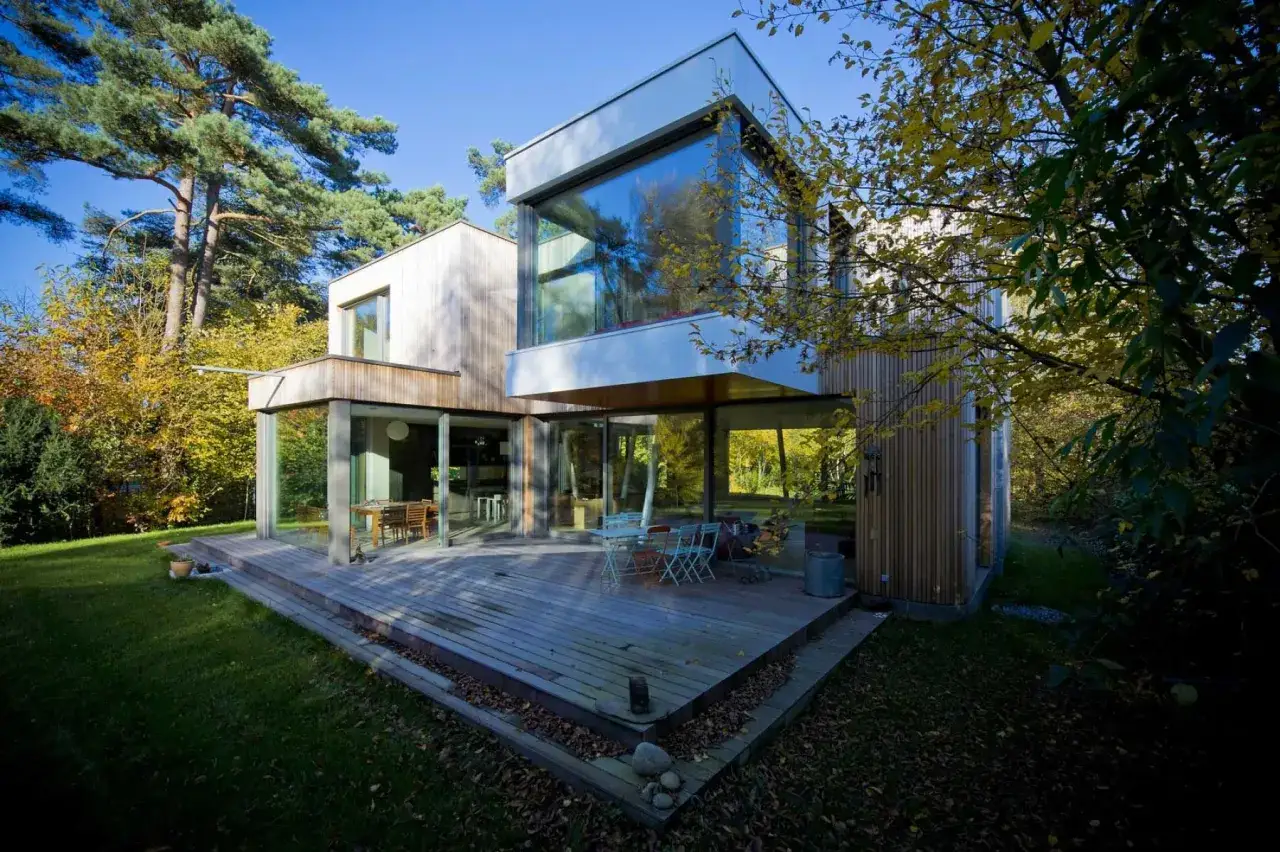 Une maison moderne simple avec une grande terrasse en bois et de larges baies vitrées, entourée d'arbres aux couleurs d'automne.