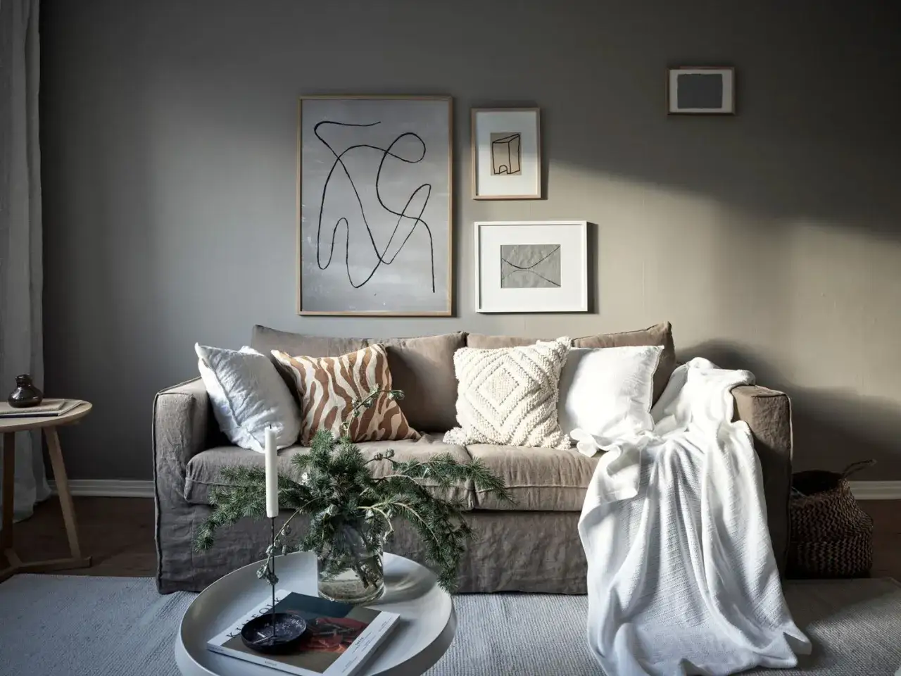 Salon cosy avec un canapé gris clair orné de coussins texturés et d'une couverture blanche. Des œuvres d'art abstraites décorent le mur.