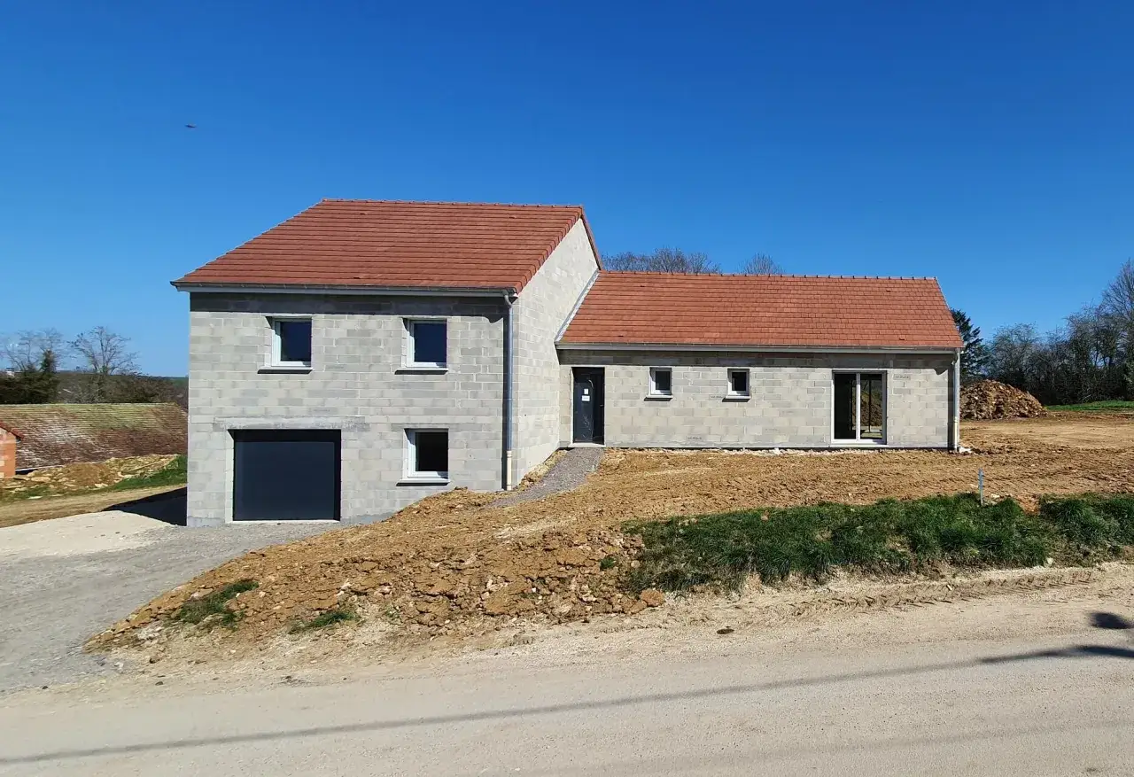 Maison prêt à finir, prix attractif. Bâtiment en parpaings gris, toit tuiles rouges, garage, jardin en friche.