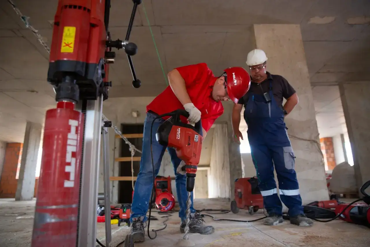 Deux ouvriers travaillent sur un chantier de pret construction neuve. L'un utilise une perceuse à percussion, l'autre supervise.