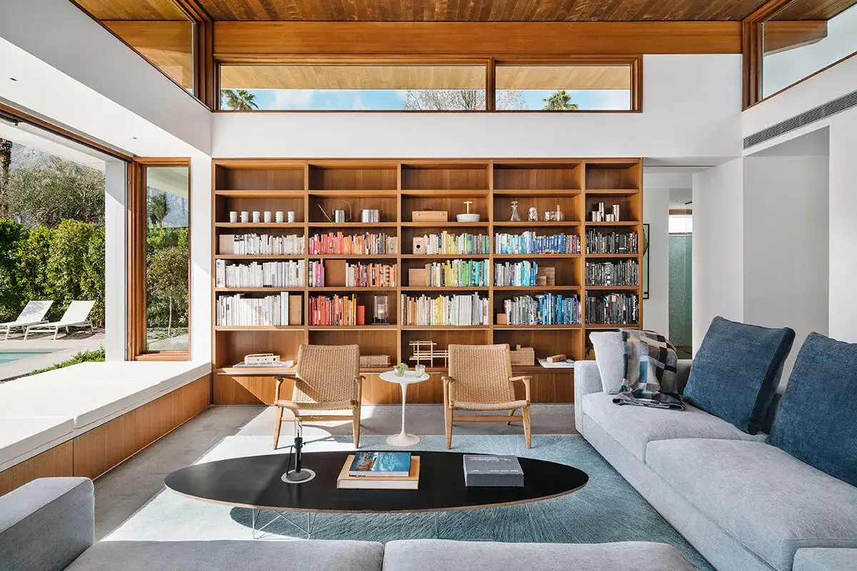 Salon lumineux avec immense bibliothèque en bois, deux fauteuils en rotin, un canapé gris et une table basse ovale. Un havre de paix, la maison bienveillante.