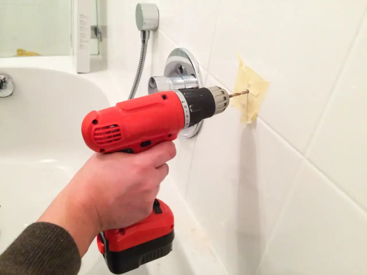 Une main utilise une perceuse pour percer du carrelage blanc, avec du ruban adhésif pour guider le foret.