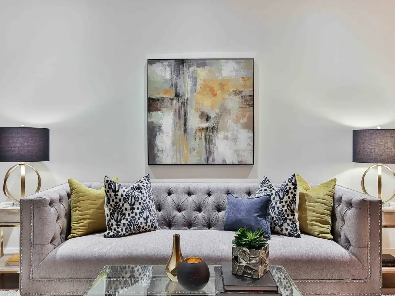 Idée pour comment décorer un grand mur de salon : un tableau abstrait moderne au-dessus d'un canapé gris capitonné, flanqué de lampes et coussins colorés.