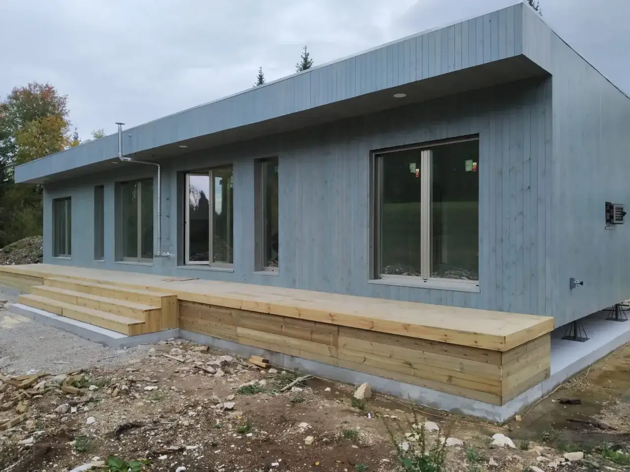 Maison modulaire moderne en bois bleu avec terrasse en bois brut et escaliers. Le chantier est encore visible.