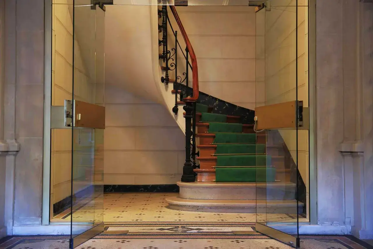 Cage d'escalier d'immeuble en r&eacute;novation, avec tapis vert, rampe en bois et balustrade en fer forg&eacute;.