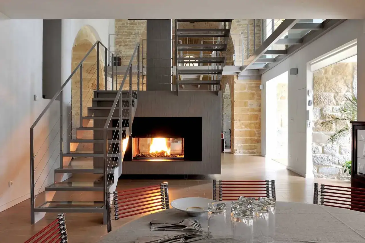 Ambiance chaleureuse dans une maison ancienne rénovée. Un feu crépite dans la cheminée centrale, flanquée d'escaliers modernes. Le plan maison ancienne révèle un espace de vie lumineux.