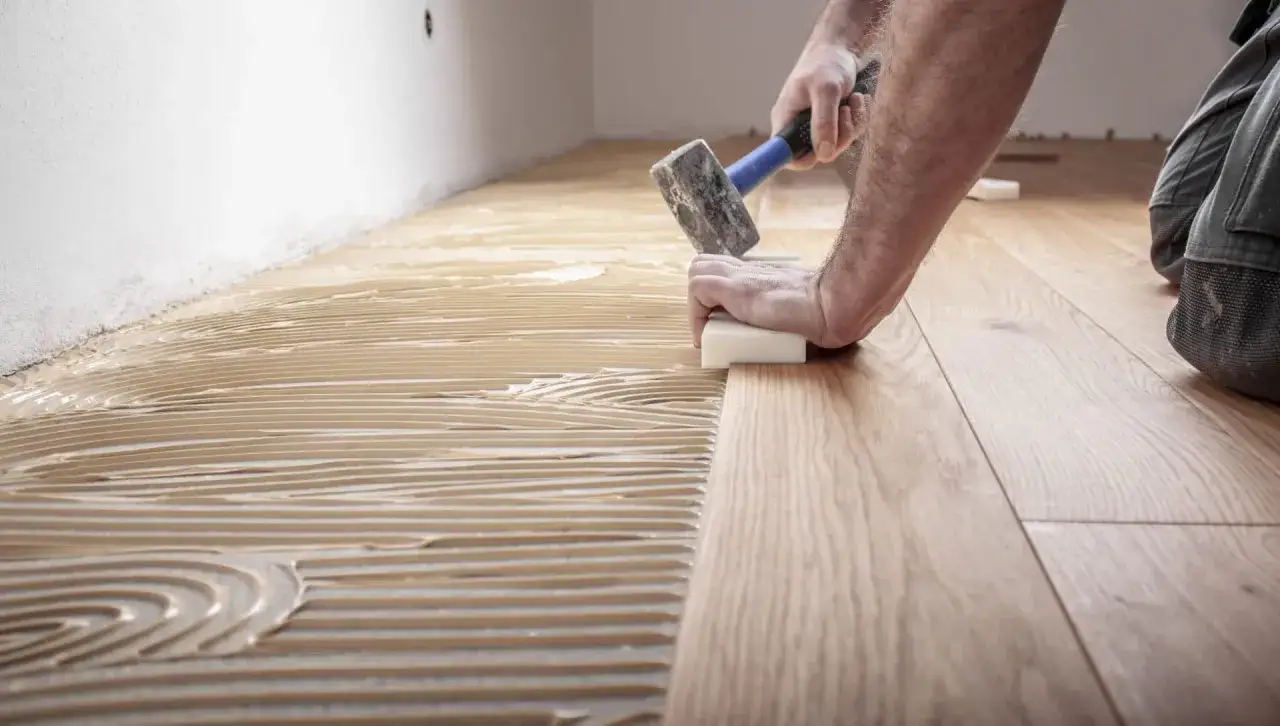 Pose d'un parquet en bois, avec de la colle appliquée au sol. Un artisan utilise un maillet pour ajuster les lames. Travaux pour refaire sol maison.