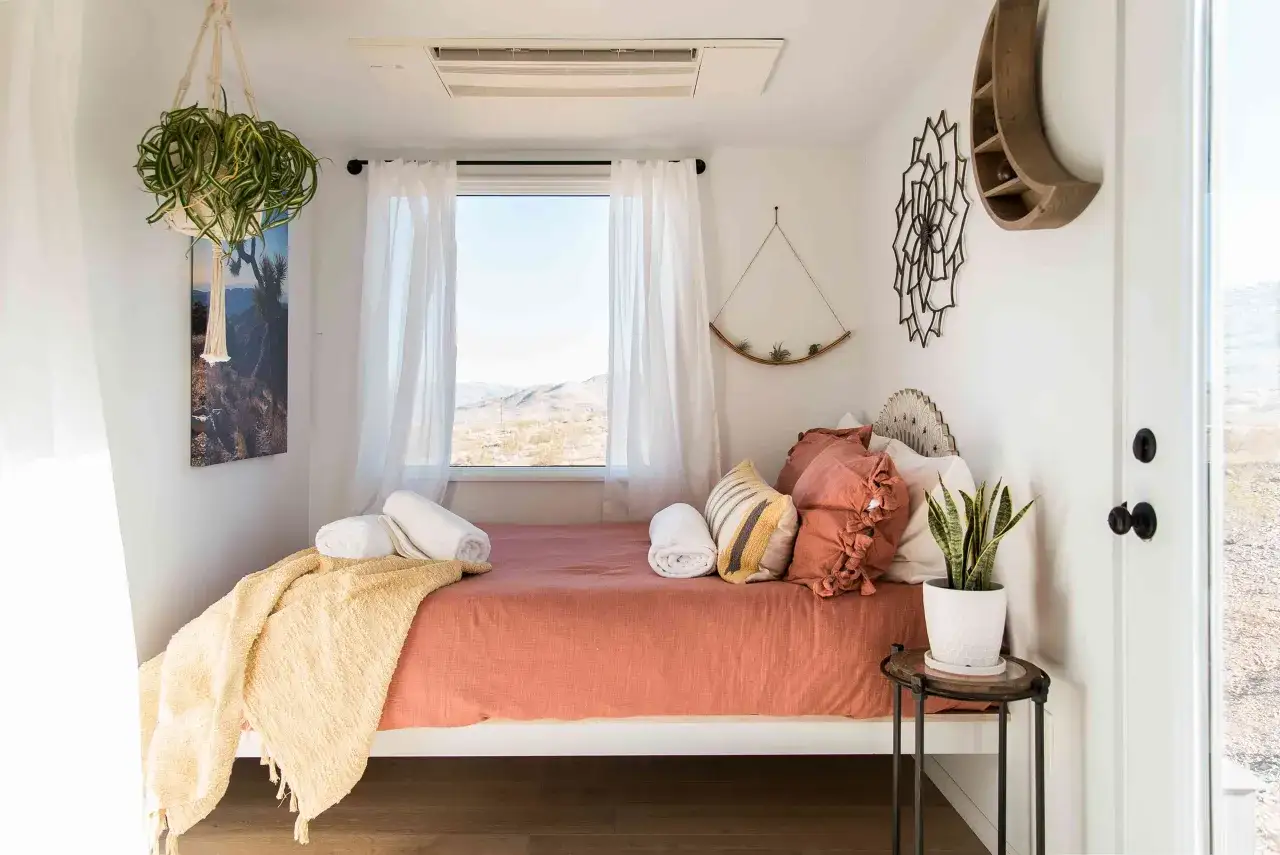 Une chambre d'amis cosy avec un lit orange, des coussins texturés, une couverture jaune et une vue désertique. Une plante suspendue ajoute une touche de verdure.