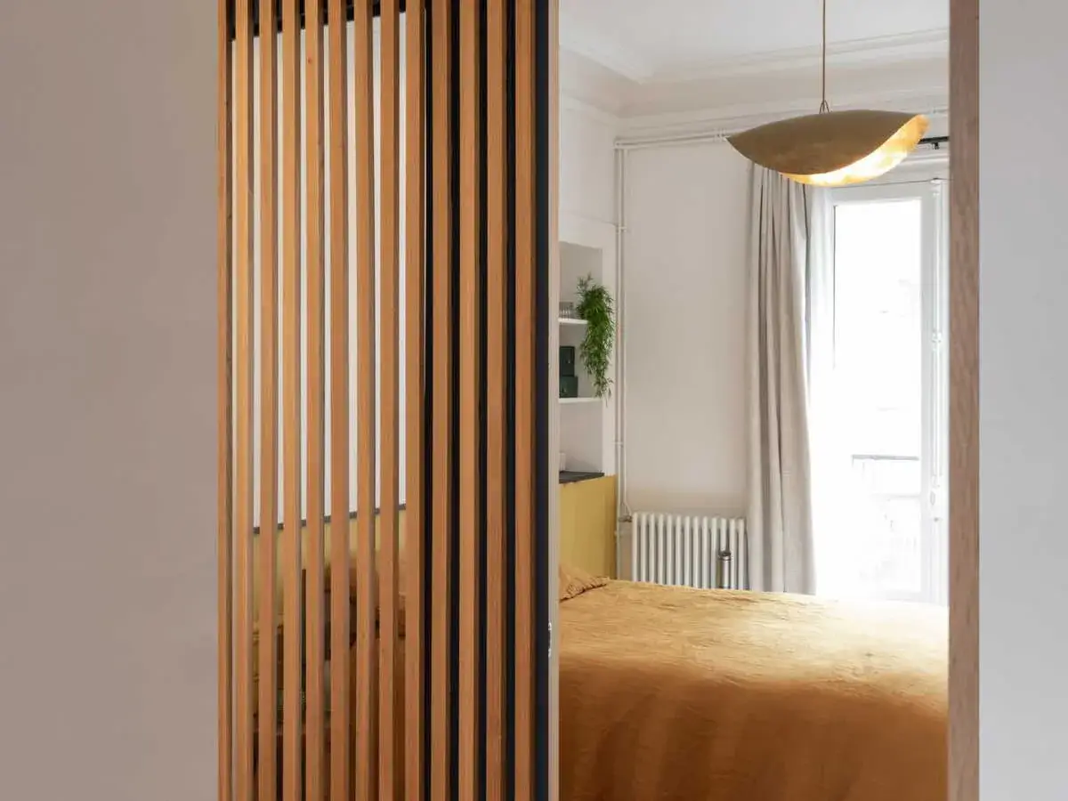 Un s&eacute;parateur de chambre en bois cr&eacute;e une division &eacute;l&eacute;gante, r&eacute;v&eacute;lant un lit douillet et une fen&ecirc;tre lumineuse.