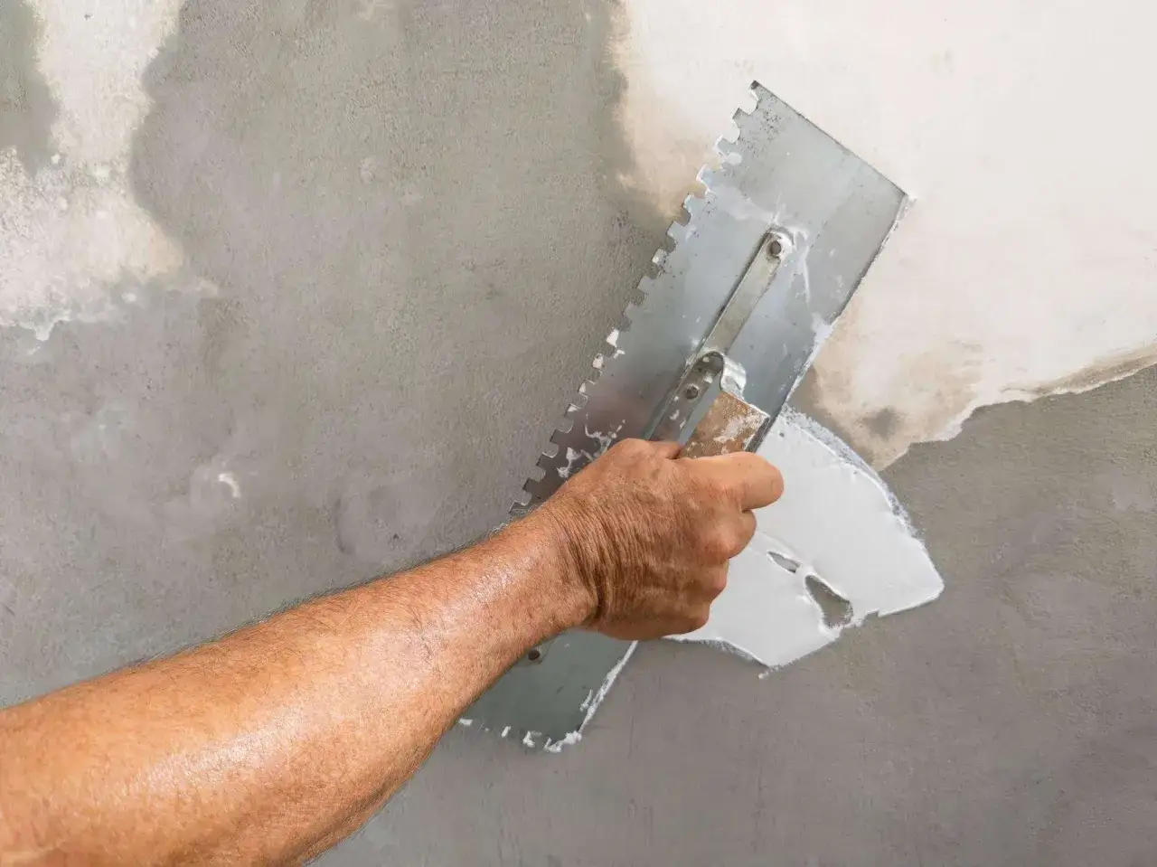 Main d'un artisan appliquant de l'enduit blanc avec une truelle dentelée pour enduire un mur abîmé.
