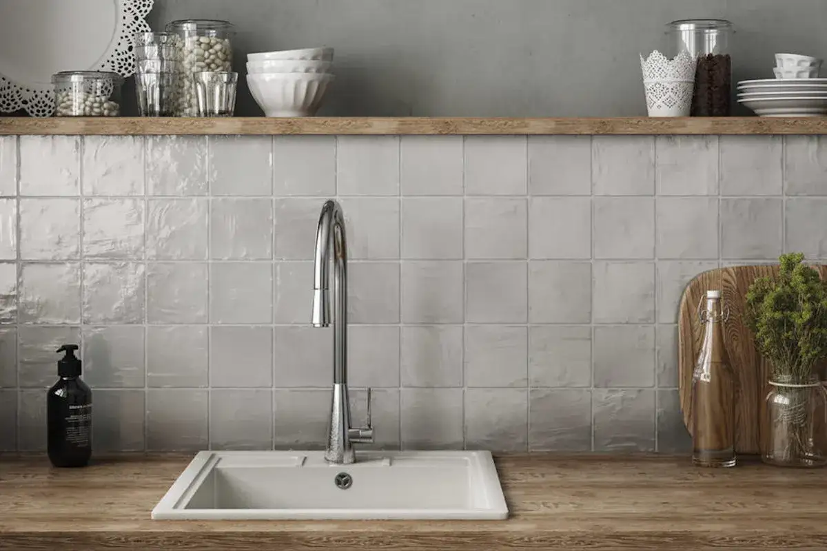 Cr&eacute;dence de cuisine avec carrelage gris clair et joints discrets. Un &eacute;vier blanc et un robinet chrom&eacute; sont visibles, ainsi que des bocaux sur une &eacute;tag&egrave;re en bois.