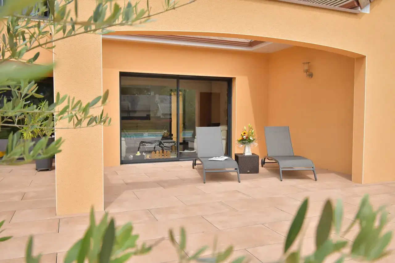 Terrasse ensoleillée d'une des maisons du midi, avec chaises longues, bouquet de fleurs et piscine visible à travers la baie vitrée.