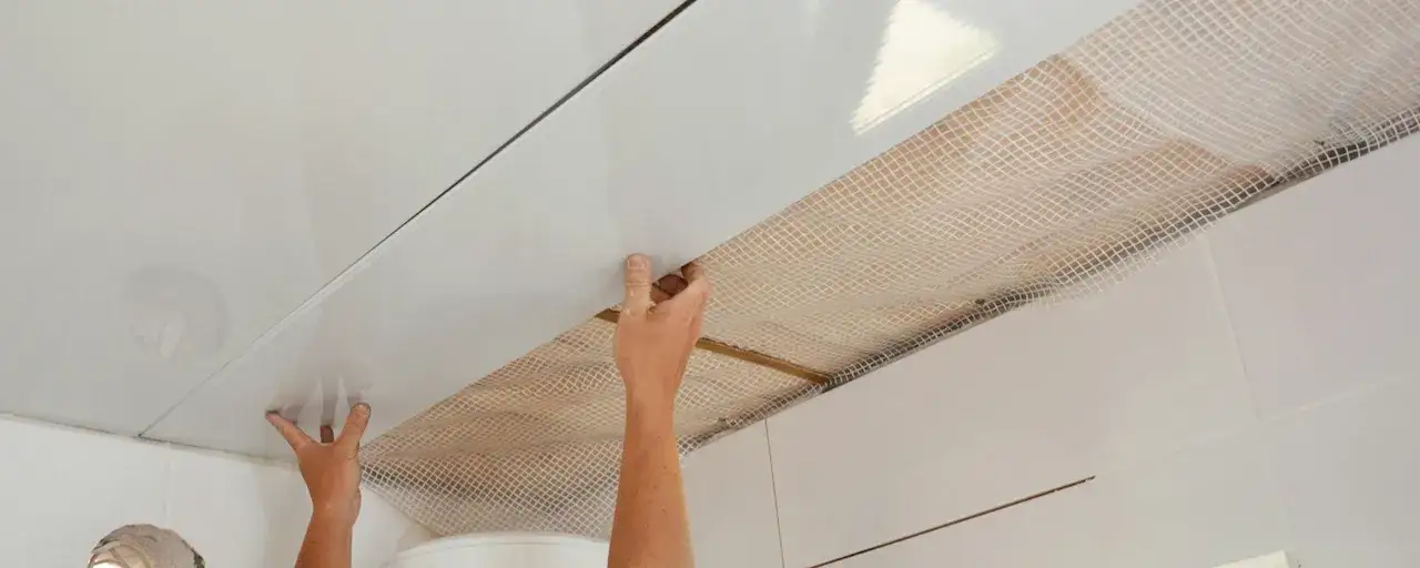 Mains installant des panneaux blancs pour refaire un plafond, avec un treillis visible en dessous.