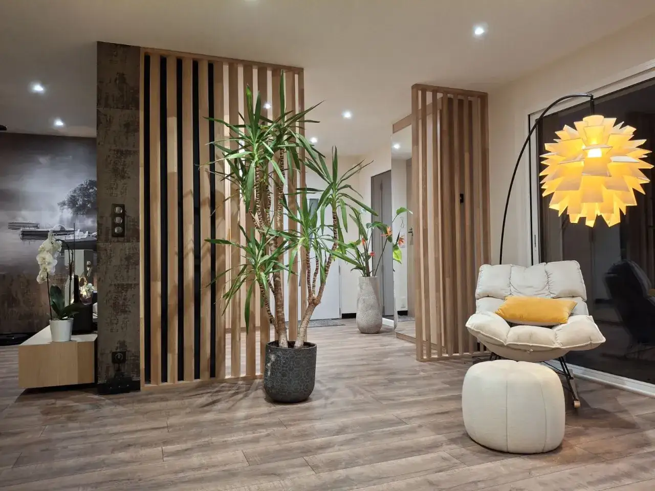 Une entrée dans un salon moderne avec un paravent en bois, une grande plante verte, un fauteuil confortable et un lampadaire design.