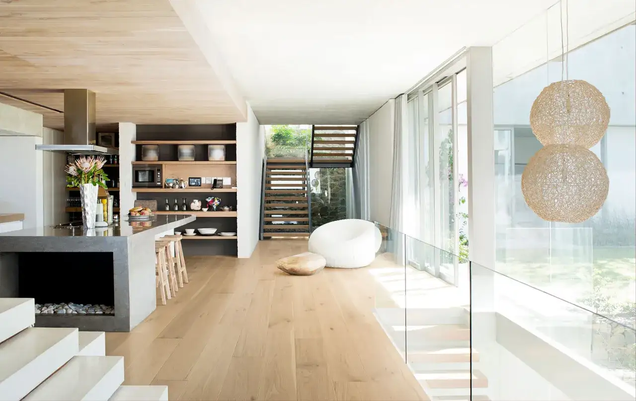 Un salon moderne avec un sol écologique en bois clair, une cuisine ouverte, des escaliers et de grandes baies vitrées.