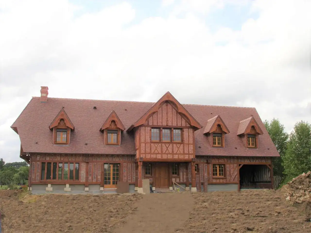 Belle maison normande en colombage, avec un toit d'ardoises rouges et de grandes fenêtres. Le jardin est en cours d'aménagement.