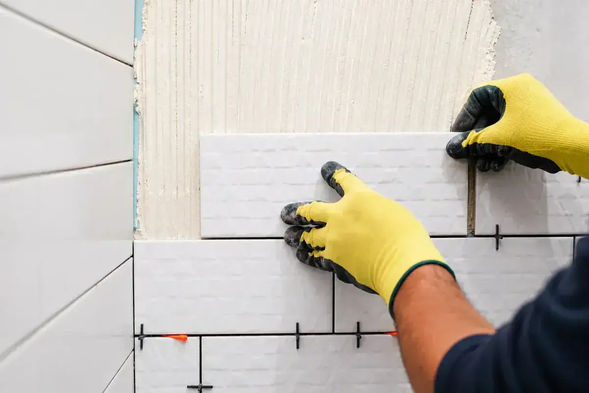 Pose de carrelage blanc, montrant l'épaisseur du carrelage et de la colle appliquée sur le mur. Des croisillons maintiennent l'espacement.