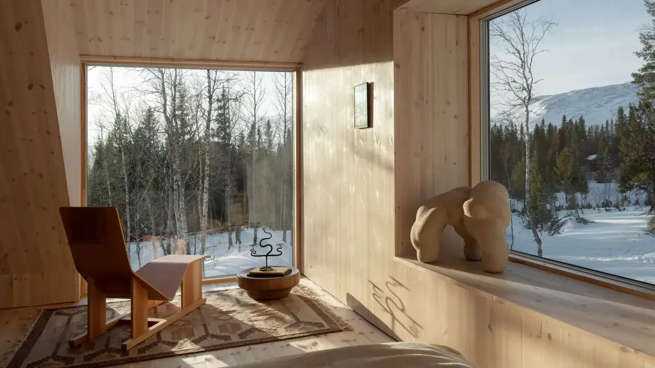 Intérieur d'une maison dans les bois, avec de grandes fenêtres offrant une vue sur une forêt enneigée et des montagnes. Un fauteuil design et une sculpture abstraite complètent le décor.