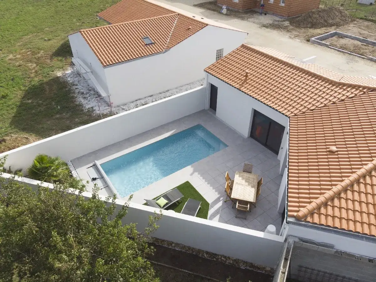 Vue aérienne d'une maison pavillonnaire avec piscine, terrasse et mobilier de jardin. Le soleil brille sur la piscine bleue et la terrasse carrelée.