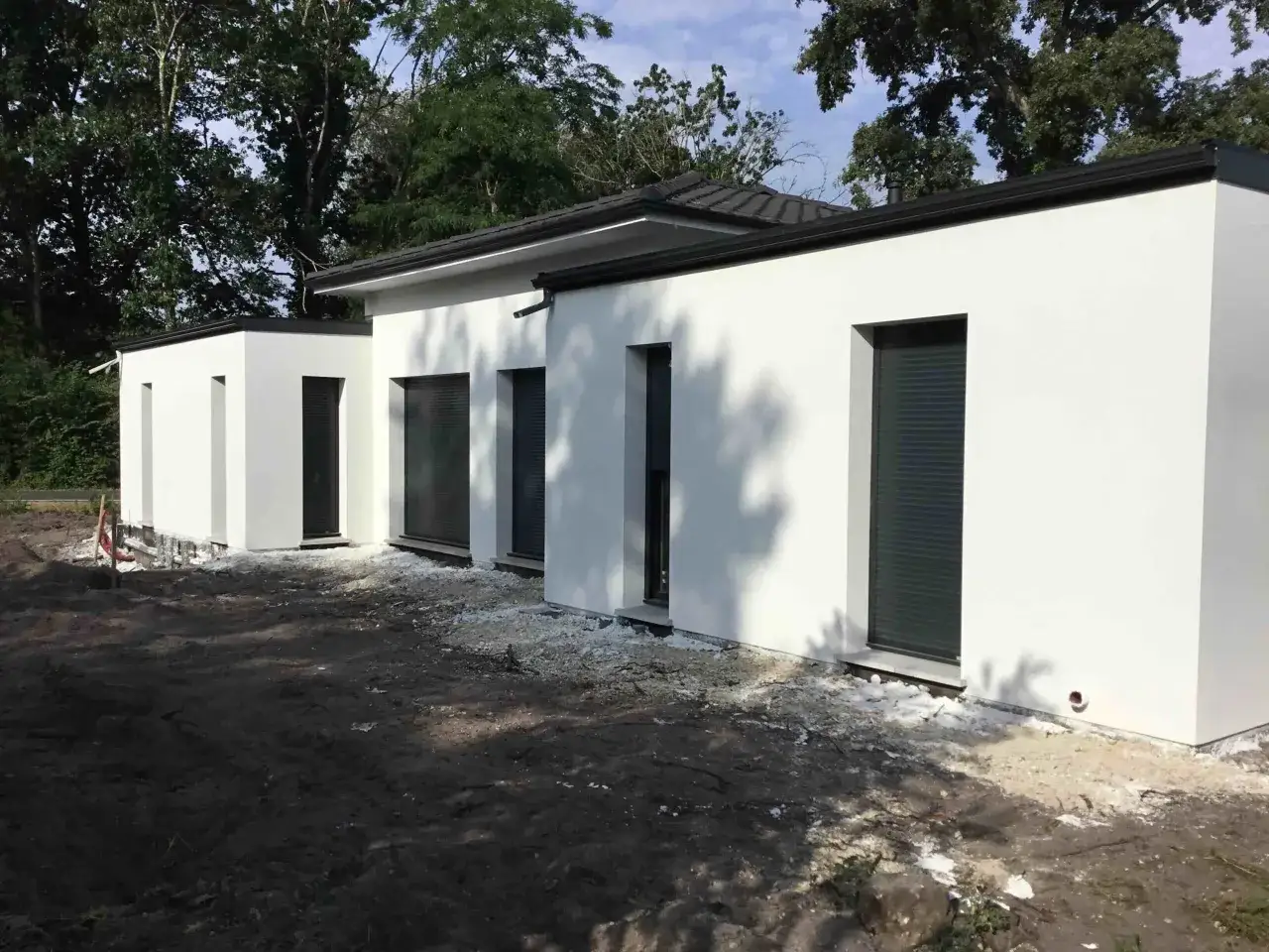 Maison moderne au style épuré, murs blancs, toit plat, fenêtres avec volets roulants sombres. Le terrain est en cours d'aménagement.