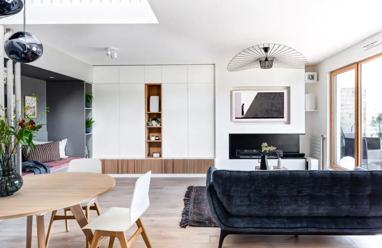 Un salon moderne et lumineux avec une table en bois, des chaises blanches, un canapé bleu nuit et une grande armoire murale. La décoration suggère un grand taille salon.