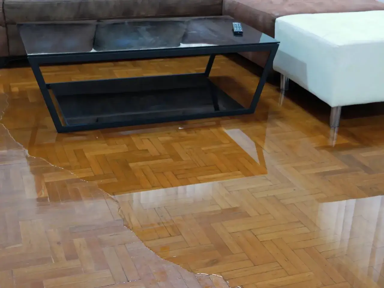 Un salon avec un parquet gondolé par l'eau. Une flaque d'eau s'étend sur le sol en bois, reflétant le mobilier.