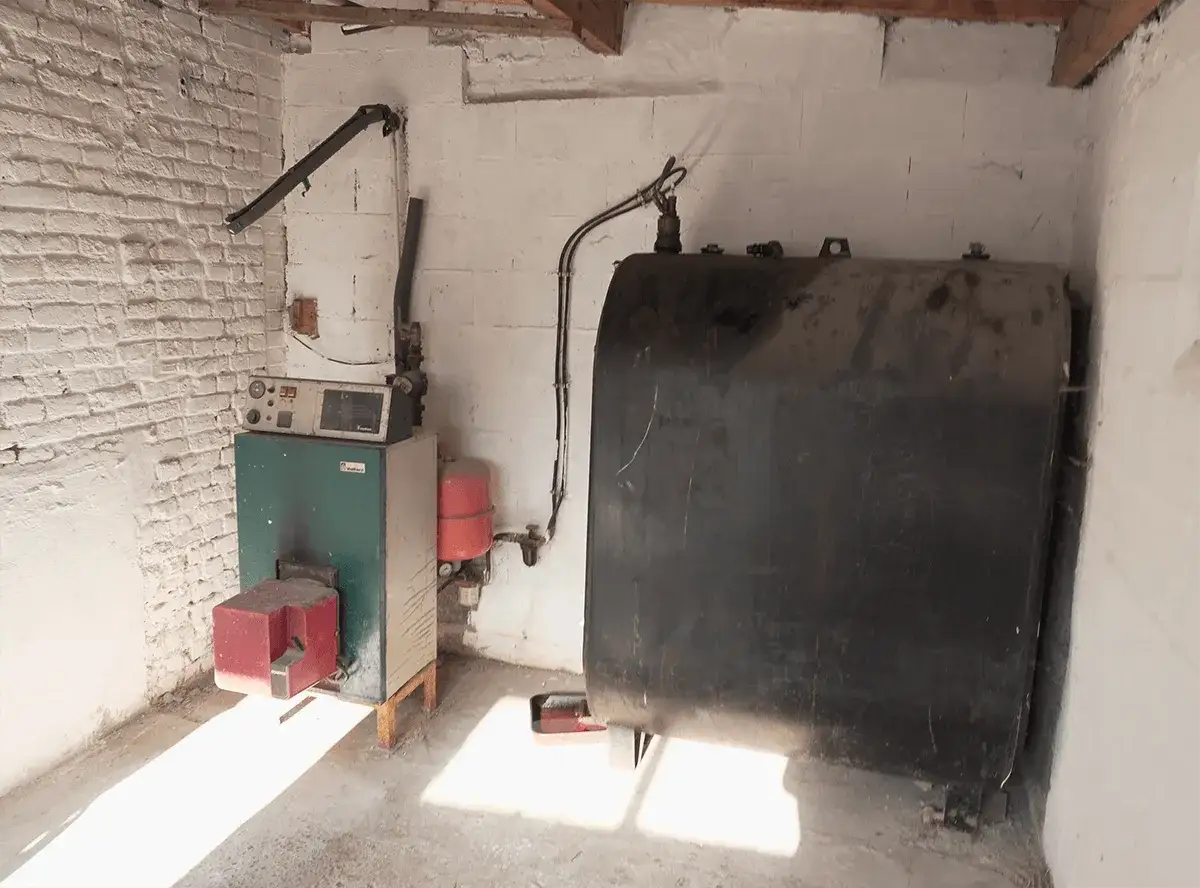 Une vieille chaudière fioul avec son réservoir noir imposant, installée dans une pièce aux murs de briques blanches.