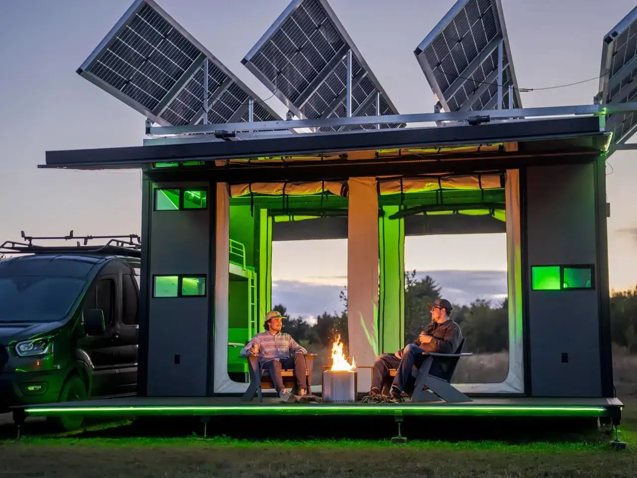 Deux hommes profitent d'un feu de camp devant une maison écologique autonome, avec des panneaux solaires sur le toit.